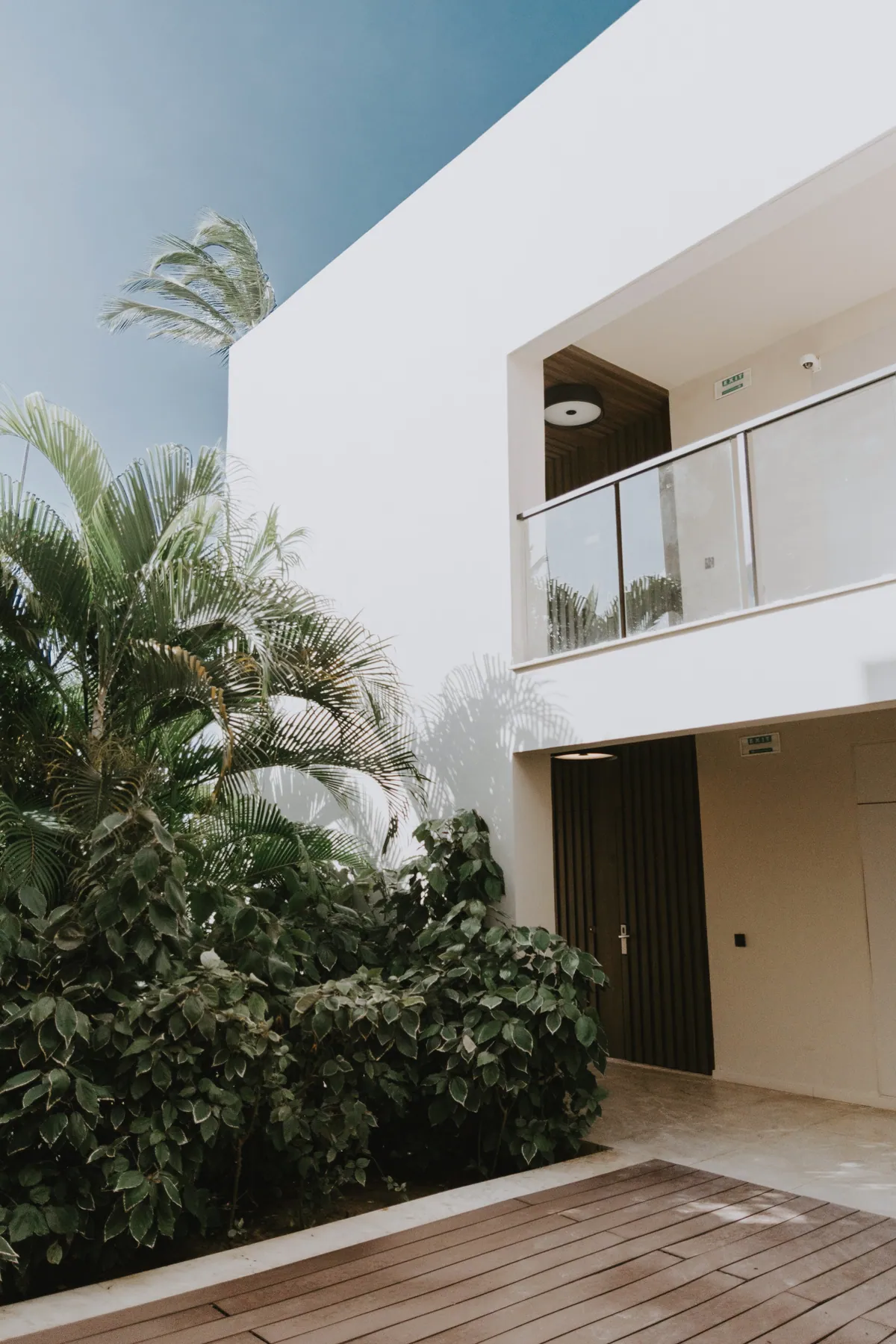 A white building with a glass-fronted balcony above a timber deck and dense tropical planting