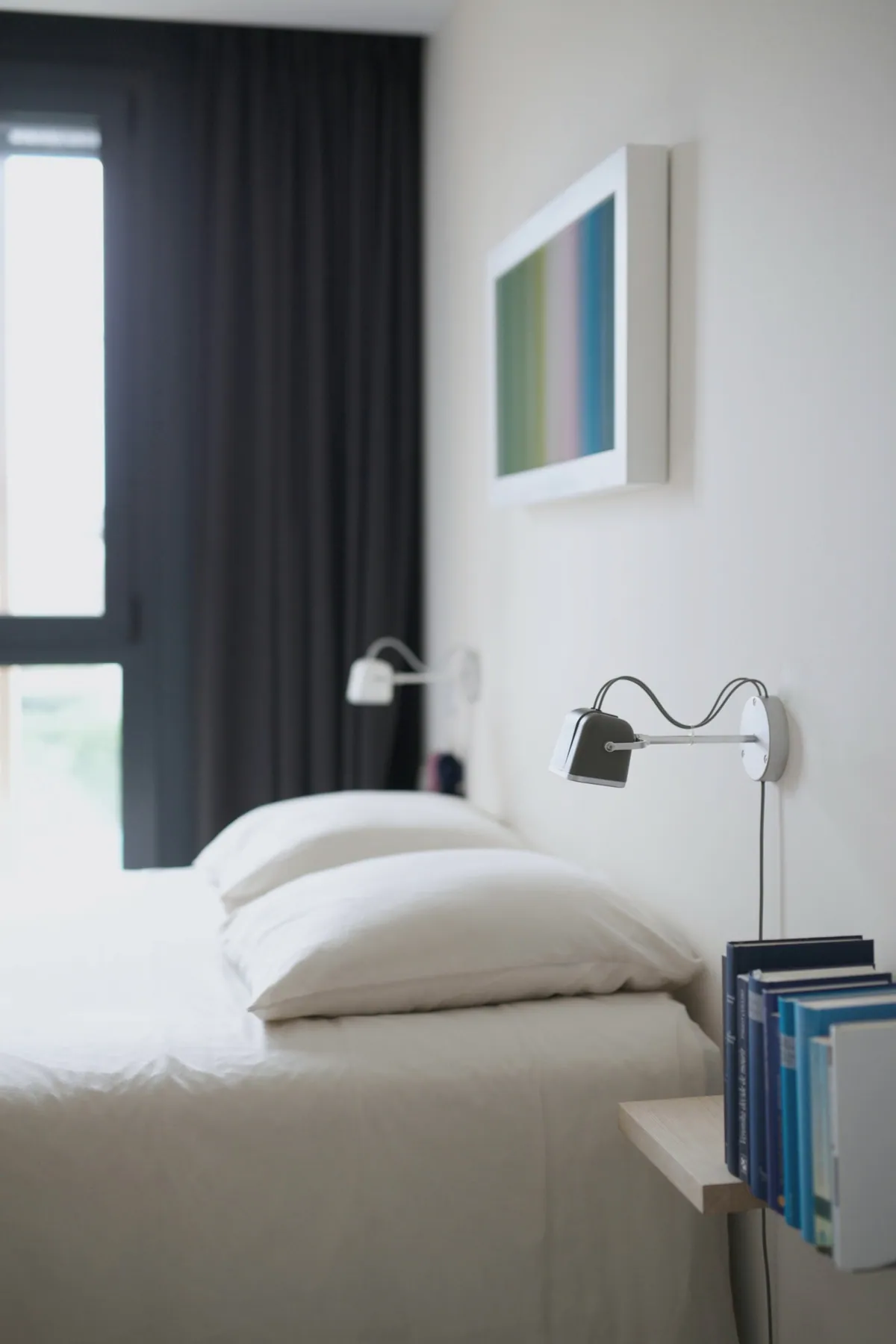 A made bed in white linen beside a tall window and a reading lamp on the wall