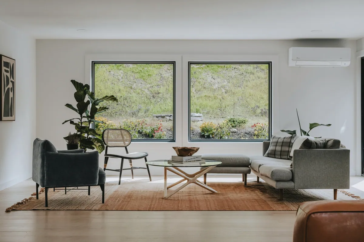 A bright living room with two large windows onto a green slope, a sofa, an armchair and indoor plants